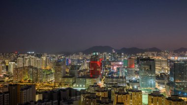 Colorful illuminated skyscrapers rise above harbourfront and distant mountains Feb 15 2026