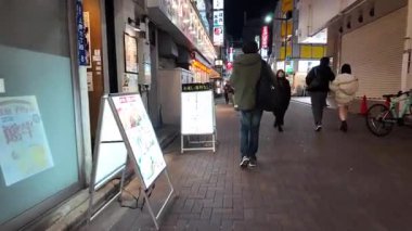 İnsanlar 9 Aralık 2026 'da kalabalık bir şehir kaldırımında parlayan menü işaretlerinin yanından geçtiler.