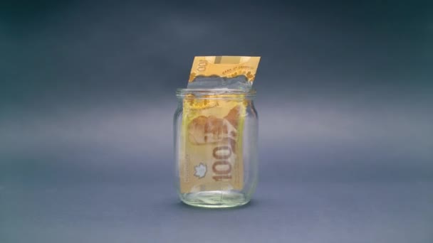 Женщина кладет 100 канадских долларов в стеклянную банку — стоковое видео