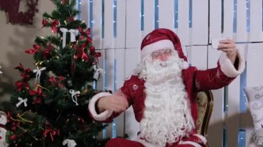 Noel Baba olduğunu oturmak ve onun Smartphone, Şömineli Oda ve Noel ağacı, hediyeler Selfi yapmak.