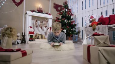 Küçük çocuk yerde yatan ve bir dijital tablet ile oynadı, kamera çocuk ve sonra çocuk, Şömine ve Noel Ağacı ile beyaz Oda hareket eder