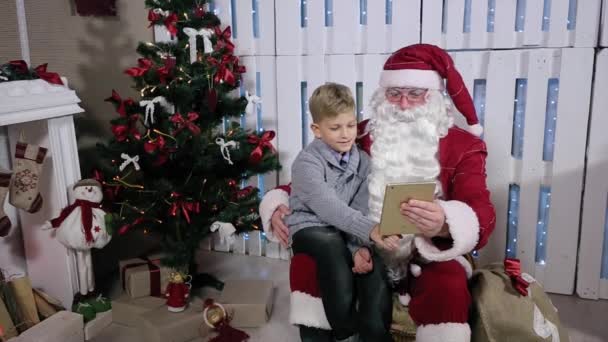 Santa och liten pojke välja presenter på tablett, sitta nära en julgran med gåvor — Stockvideo