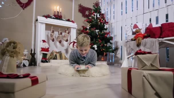 Riéndose niño jugando en la tableta digital, La cámara se mueve en un niño, Habitación blanca con chimenea y árbol de Navidad — Vídeo de stock