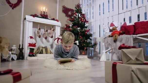 De jongen kiest een gift voor de digitale tablet, de camera wordt verplaatst naar een jongen, beweegt de camera van jongen, witte kamer met open haard en kerstboom — Stockvideo