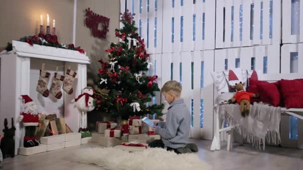 Niño utilizando la tecnología de tableta táctil, convierte la tableta digital en las manos de verlo, Habitación blanca con chimenea y árbol de Navidad — Vídeo de stock