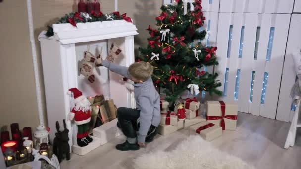 Een jongen op zoek geschenken tenen bij de open haard, witte kamer met open haard en kerstboom. Groothoek — Stockvideo