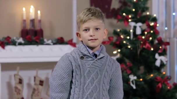 Menino caucasiano expressivo feliz mostrando na câmera o sinal polegar para cima no estúdio de Natal, sorrindo, sala branca com lareira e árvore de Natal, Médio — Vídeo de Stock