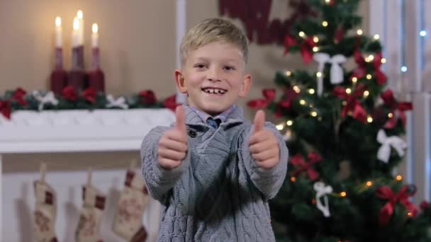 Μικρό αγόρι που κρατά ψηλά δύο δάχτυλα, το χαμόγελο, βλέμμα φωτογραφική μηχανή, λευκό δωμάτιο με τζάκι και χριστουγεννιάτικο δέντρο, μέσο — Αρχείο Βίντεο