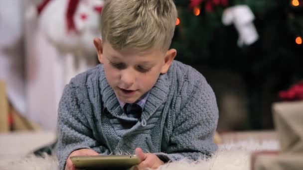 Маленький хлопчик лежав на землі і грав з цифровим планшетом, камера рухається на хлопчика, білий кімнату з каміном і Різдвяна ялинка, крупним планом, люди — стокове відео