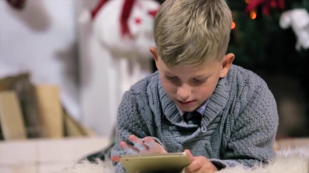 Ragazzino sdraiato a terra e giocato con un tablet digitale, La fotocamera si muove dal ragazzo e poi il ragazzo, camera bianca con camino e albero di Natale, Close Up, persone — Video Stock