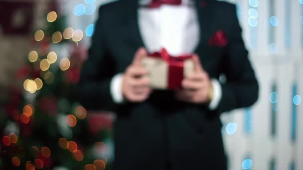 Человек в черном костюме дарит зрителю подарок с красной лентой в белой комнате с камином и елкой — стоковое видео