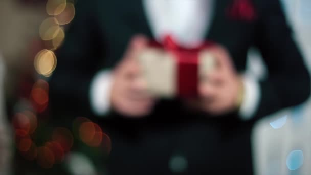 En man i svart dräkt ger en gåva till betraktaren, ljuset av julgran — Stockvideo