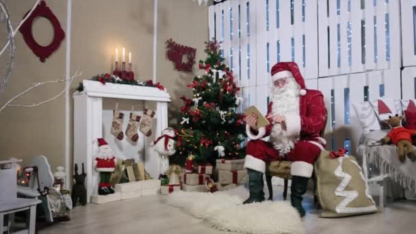 Santa da la vuelta a la página en la sala de la tableta con chimenea y regalos del árbol de Navidad . — Vídeos de Stock