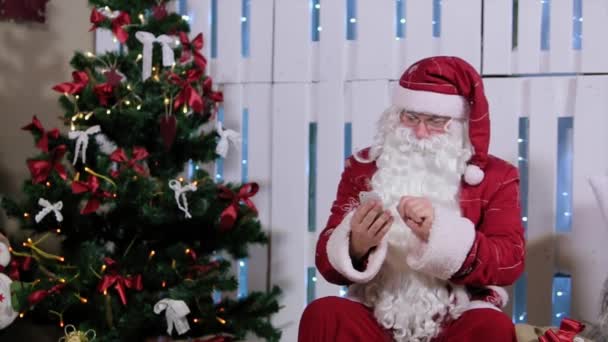 Santa Claus está sentado y hablando su teléfono, habitación con chimenea y árbol de Navidad, regalos . — Vídeos de Stock