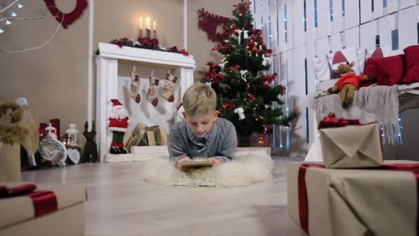 Lachender Junge spielt auf digitalem Tablet, die Kamera fährt auf einen Jungen, weißes Zimmer mit Kamin und Weihnachtsbaum — Stockvideo