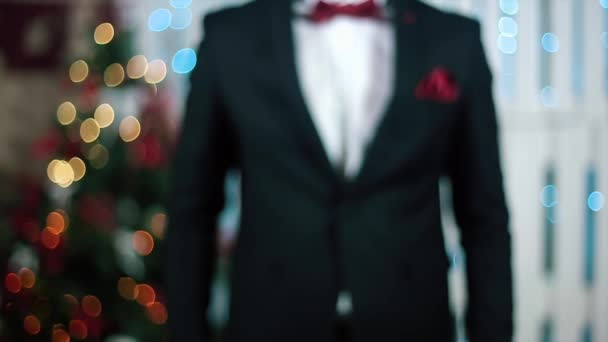 Man i svart kostym ger kameran en gåva med ett rött band i ett vitt rum med öppen spis och julgran — Stockvideo