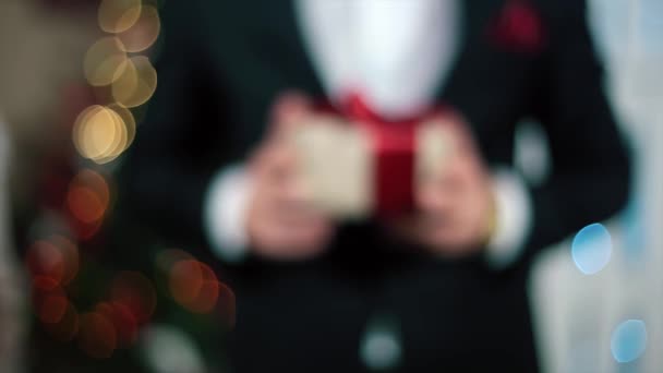 Człowiek w czarnym garniturze daje prezent dla widza, w świetle choinki — Wideo stockowe
