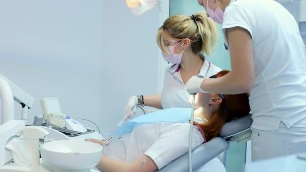 Un dentiste masqué et ganté explique le traitement à venir à sa patiente, puis commence l'examen, à l'aide de sondes dentaires, aidé par son assistante dentaire avec un tube d'aspiration — Video