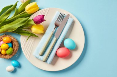 Paskalya sofrasında laleler, pastel yumurtalar ve mavi arka planda tabaklar.