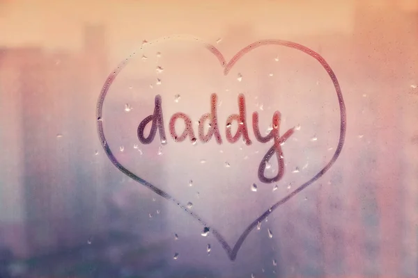 Handwritten word Daddy in heart shape on misted sunset pink window with raindrops