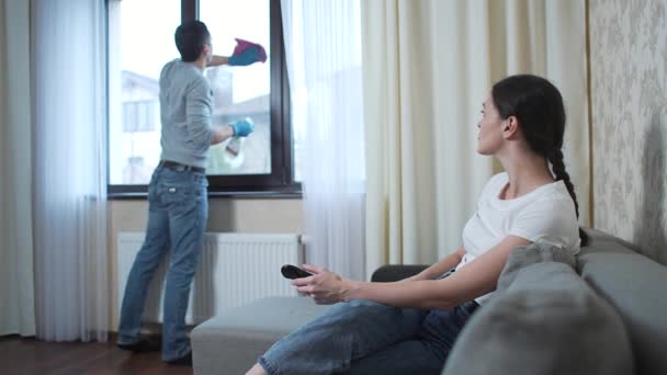 Mari lave la fenêtre et femme regarde la télévision — Video