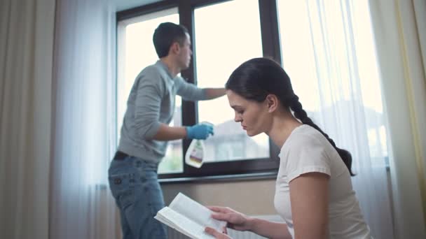 Esposo limpia la ventana y esposa leyendo el libro — Vídeo de stock