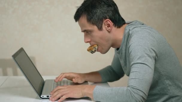 Человек с кексом во рту, работающий над ноутбуком — стоковое видео