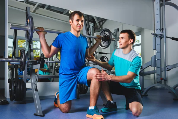 Esporte, fitness, trabalho em equipe, musculação conceito de pessoas homem e personal trainer com músculos de flexão barbell no ginásio — Fotografia de Stock