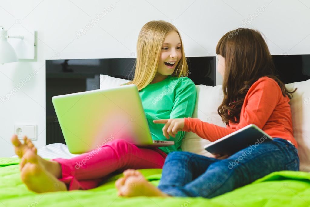 Two beautiful little sisters sitting on bed and play with a