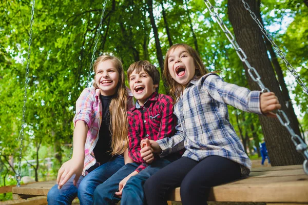 Bambini sorridenti che si divertono al parco giochi. Bambini che giocano all'aperto in estate. Adolescenti che cavalcano su un'altalena fuori — Foto Stock