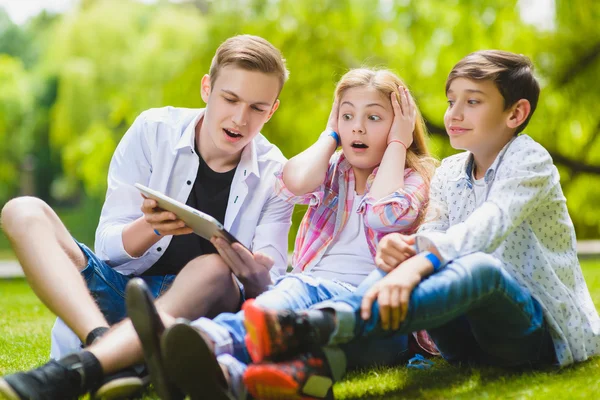 Uśmiechnięte dzieci bawią się i patrzeć na tablet w trawie. Dzieci bawią się na świeżym powietrzu w lecie. Nastolatki komunikują się na zewnątrz — Zdjęcie stockowe