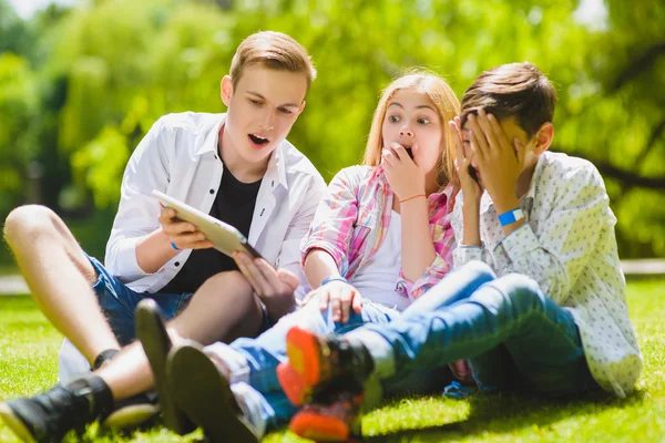 Uśmiechnięte dzieci bawią się i patrzeć na tablet w trawie. Dzieci bawią się na świeżym powietrzu w lecie. Nastolatki komunikują się na zewnątrz — Zdjęcie stockowe
