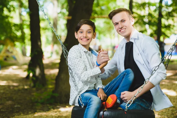 Ragazzi sorridenti che si divertono al parco giochi. Bambini che giocano all'aperto in estate. Adolescenti che cavalcano su un'altalena fuori — Foto Stock