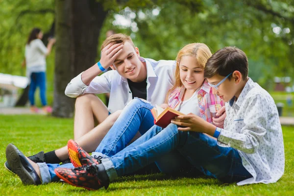 Uśmiechnięte dzieci zabawy i czytania książki na trawie. Dzieci bawią się na świeżym powietrzu w lecie. Nastolatki komunikują się na zewnątrz — Zdjęcie stockowe