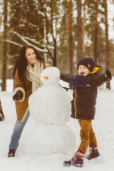 Familia feliz con ropa de abrigo. Sonriente madre e hijo haciendo un muñeco de nieve al aire libre. El concepto de actividades invernales —  Fotos de Stock