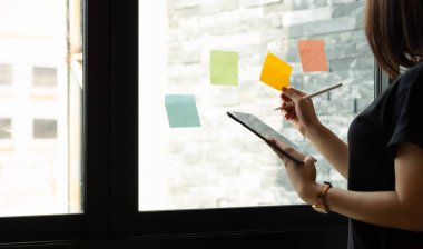 Post it sırasında tablet kullanan iş kadını toplantı odasındaki cam duvarda tartışma ve planlama fikrini not alıyor..