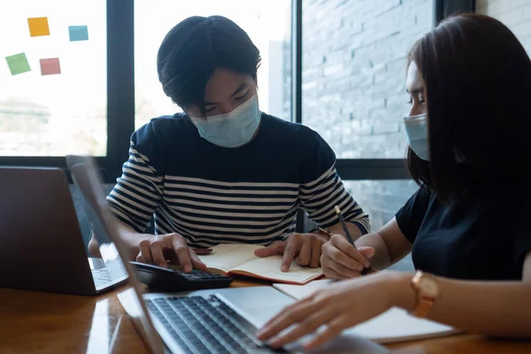 Asian woman and man workers meeting together with laptop for financial and wear protective masks prevent corona virus or covid19 at co working space .Health and teamwork concept.