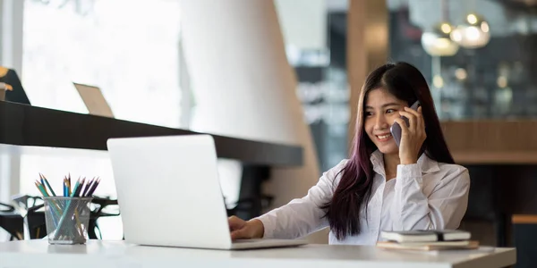 젊은 아시아 사업가 가 사무실에서 랩탑 컴퓨터로 타자를 치면서 아름다운 미소를 짓고 휴대 전화로 이야기하는 모습 — 스톡 사진