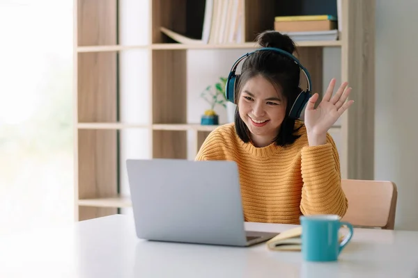 Glückliche junge asiatische Ethnizität weibliche Managerin trägt drahtlose Kopfhörer, Blick auf Laptop-Bildschirm, hält angenehme Gespräche mit Partnern Kunden online, Arbeit aus der Ferne am Arbeitsplatz. — Stockfoto