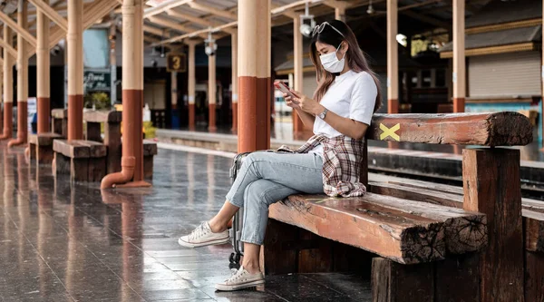 Asiático turista adolescente usando máscara esperando distância estação de trem usando smartphone, mídias sociais check-in, viajante solitário, Verão férias conceito de aventura ferroviária. — Fotografia de Stock