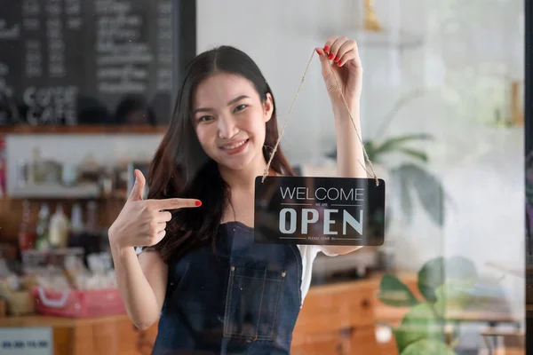 Retrato de sorridente jovem barista menina em avental segurando placa de sinal aberto enquanto estava em seu café. elegante asiático café feminino staff virar placa da porta na manhã em própria loja pequeno negócio — Fotografia de Stock