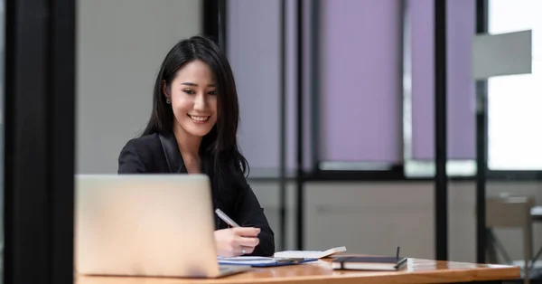 Ofisinde iş yerinde oturan mutlu Asyalı genç iş kadını. Ofiste dizüstü bilgisayarda çalışan genç bir kadın. — Stok fotoğraf