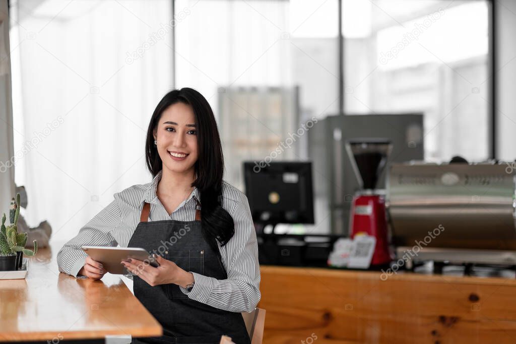 Cheerful young asian woman owner holding digital tablet while standing in her cafe, young entrepreneur conceptual