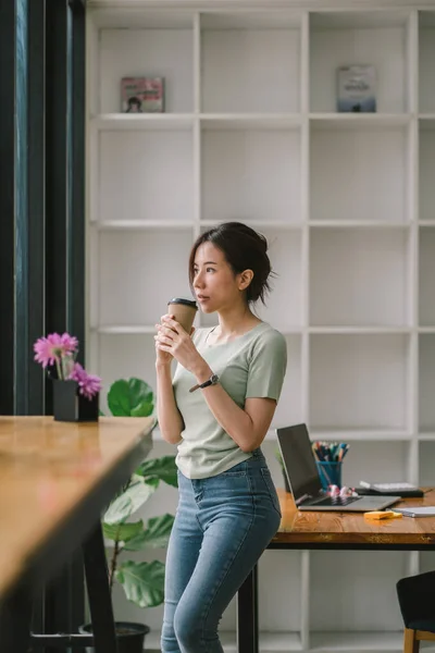 Mujer asiática parada frente al lugar de trabajo de la oficina mientras sostiene una taza de café y mira afuera — Foto de Stock