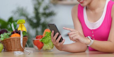 Taze sebzelerle çevrili akıllı telefonuyla etkileşime geçen bir kadın, teknolojinin sağlıklı yaşama entegrasyonunu vurguluyor..