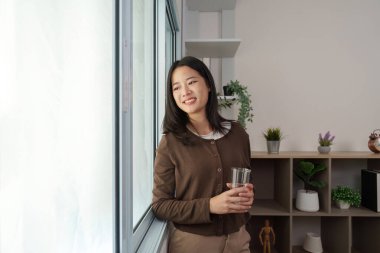 Pencerenin kenarında duran genç bir kadın bir bardak tutuyor ve gülümsüyor. Huzur ve iç gözlem anını yansıtıyor..