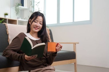 Bir kadın kanepeye oturur, kitap okur, kahvesini yudumlar, rahat bir rahatlama ve keyif ortamı yaratır..