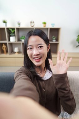 Genç bir kadın kameraya gülümser ve selfie çekerken el sallar. Neşeli bir bağlantı anı yakalar..