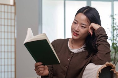 Bir kadın rahat bir şekilde oturur, düşünceli bir ifadeyle kitap okur, edebiyat ve kişisel zamanın zevkini vurgular..