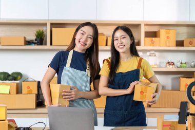 Bir e-ticaret çalışma alanında paketleri gururla sergileyen iki kadın, takım çalışması ve iş dünyasında başarı sergiliyor..